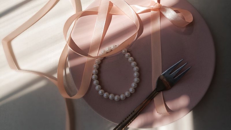 Photograph of a pearl bracelet and loose blush satin ribbon on a pale pink plate in bright window light