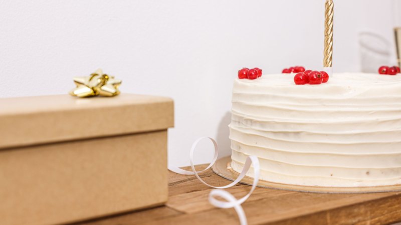 Photograph of a white frosted cake with red currants and a single gold candle beside a kraft gift box with a gold bow on a wooden shelf