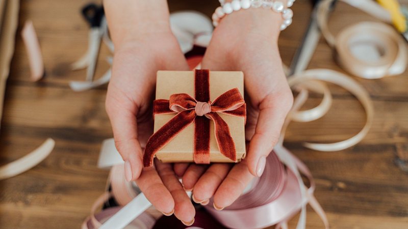 Photograph of hands holding a small kraft-paper gift box tied with a rust velvet ribbon
