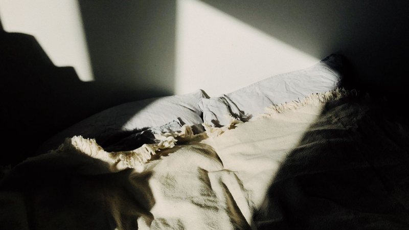 Photograph of an unmade bed with crumpled white pillows and a fringed cream throw lit by a slash of low sunlight in a dark bedroom