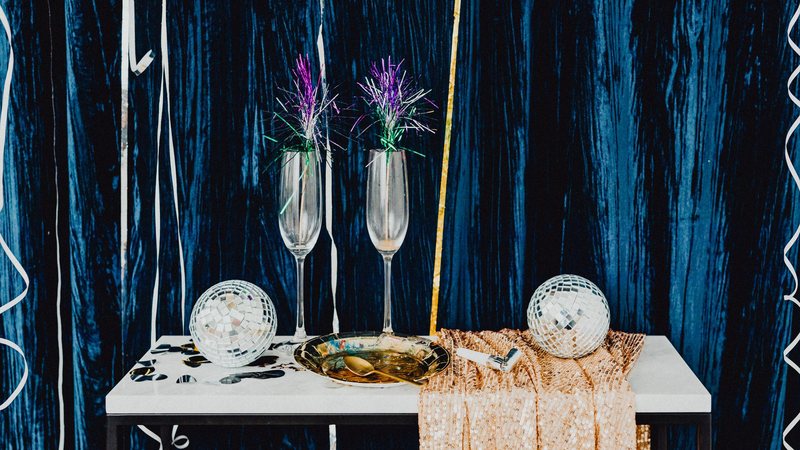 Photograph of two Champagne flutes, mirror balls and a gold tray on a white table in front of a dark blue velvet curtain